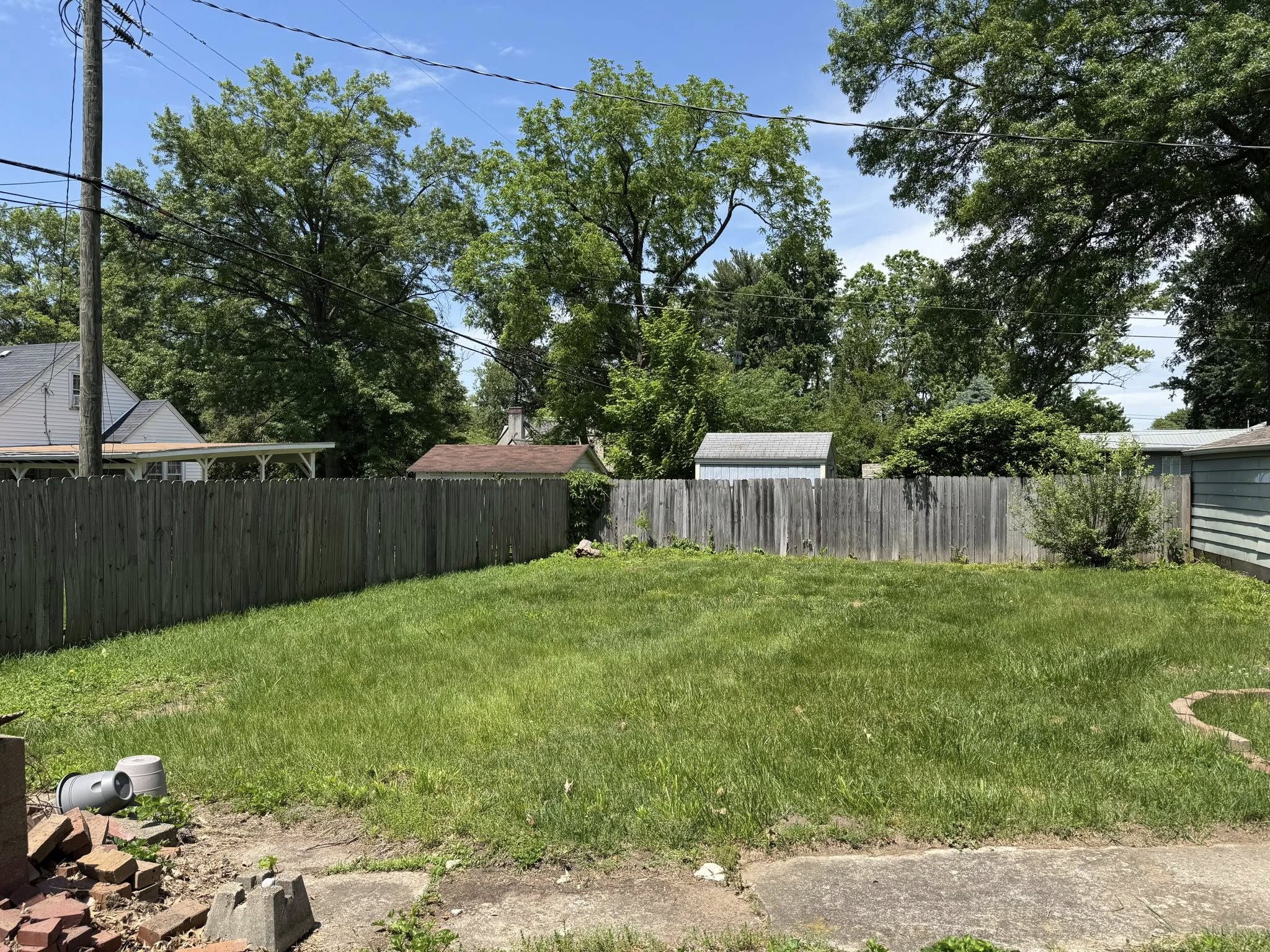 A fenced backyard with green grass, some overgrown areas, and trees in the background on a sunny day.