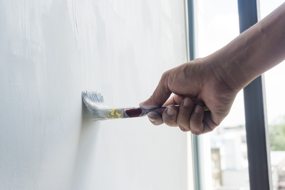 A construction company worker is painting a white wall with a paintbrush near a window.