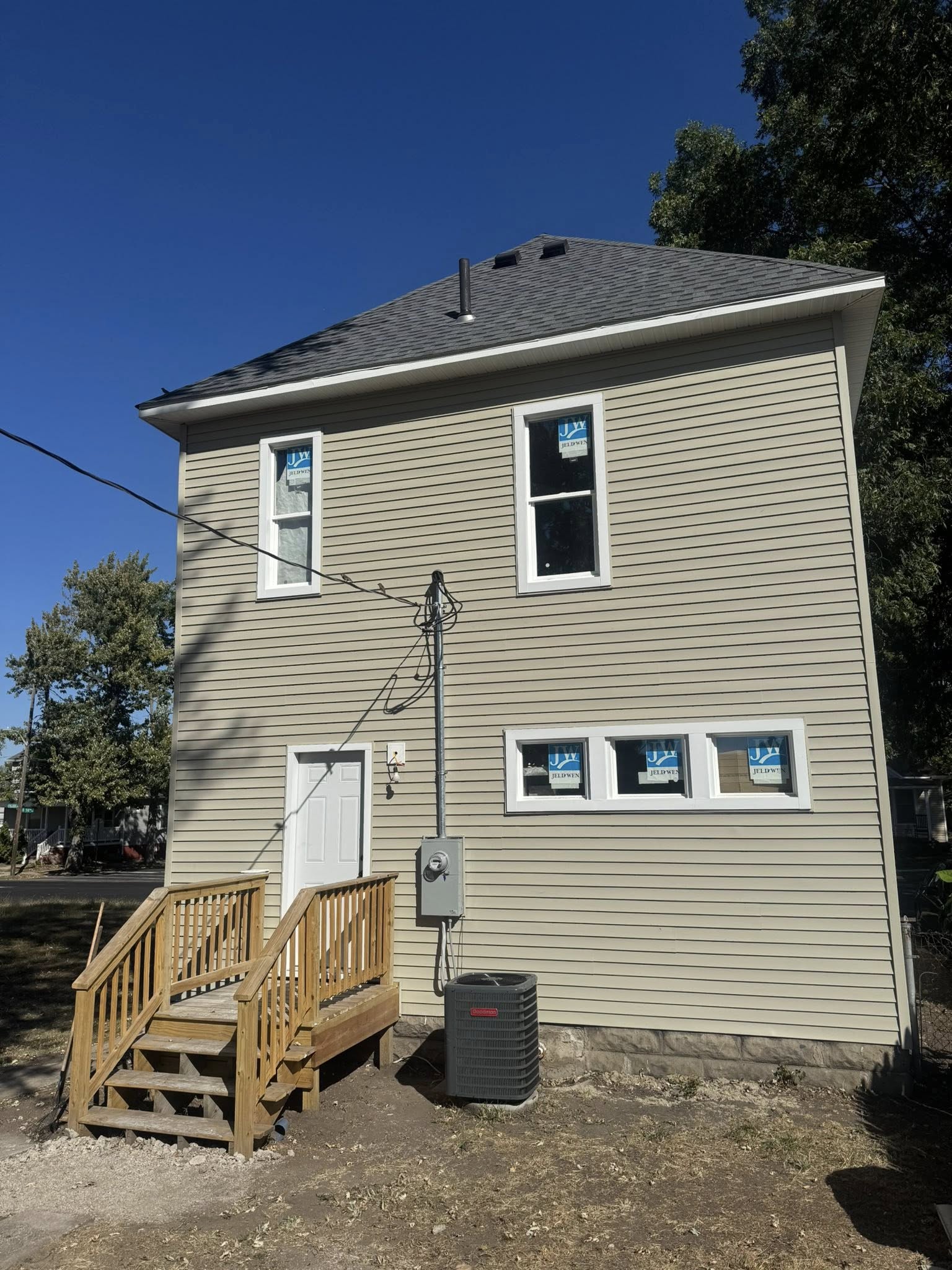A beige two-story house with new siding by a trusted construction company, several windows with blue stickers, a small wooden porch, and an outdoor HVAC unit under a clear sky.