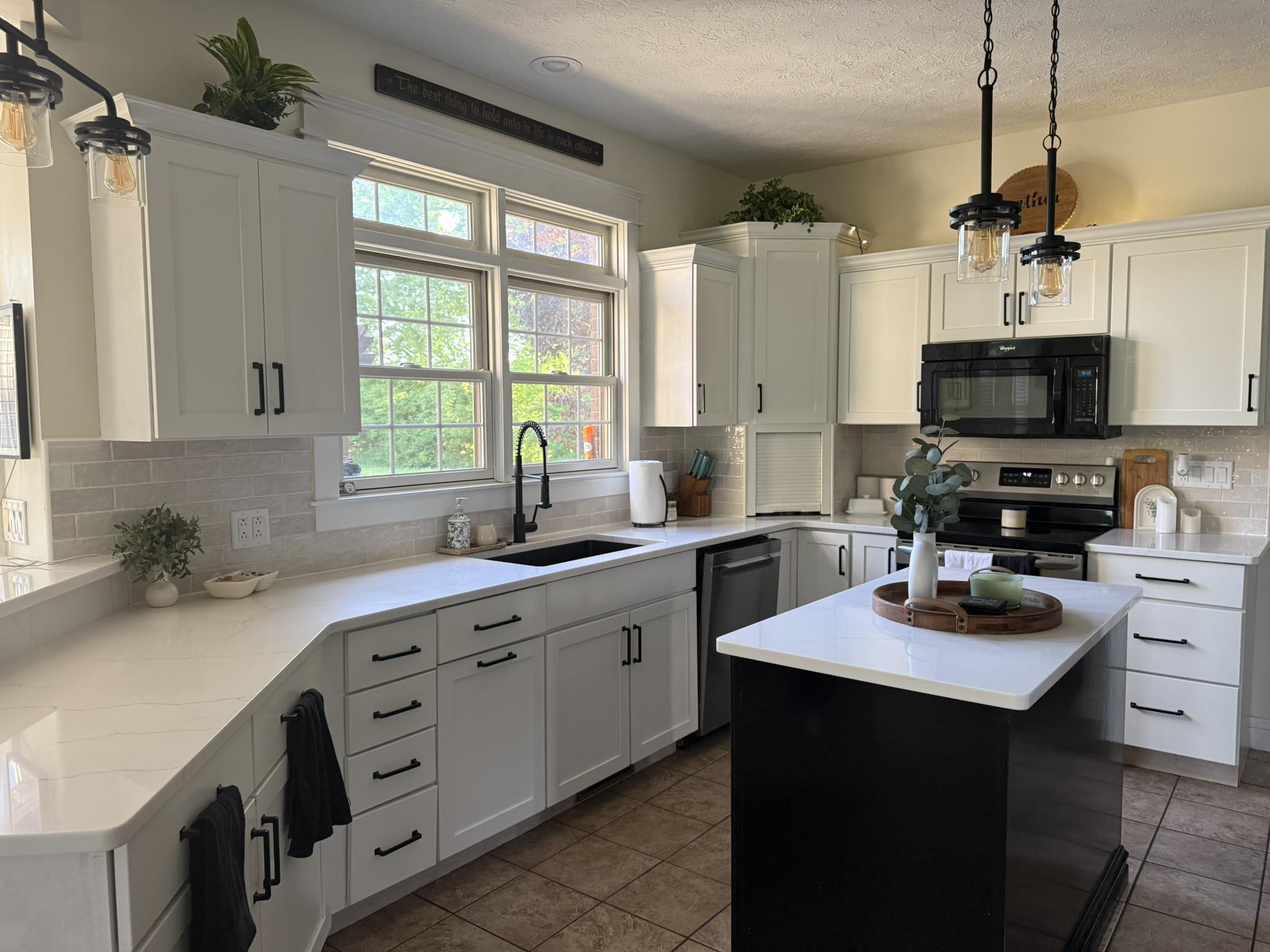 Modern kitchen with white cabinets, black hardware, an island, stainless steel oven, black microwave, farmhouse sink, and large windows letting in natural light.
