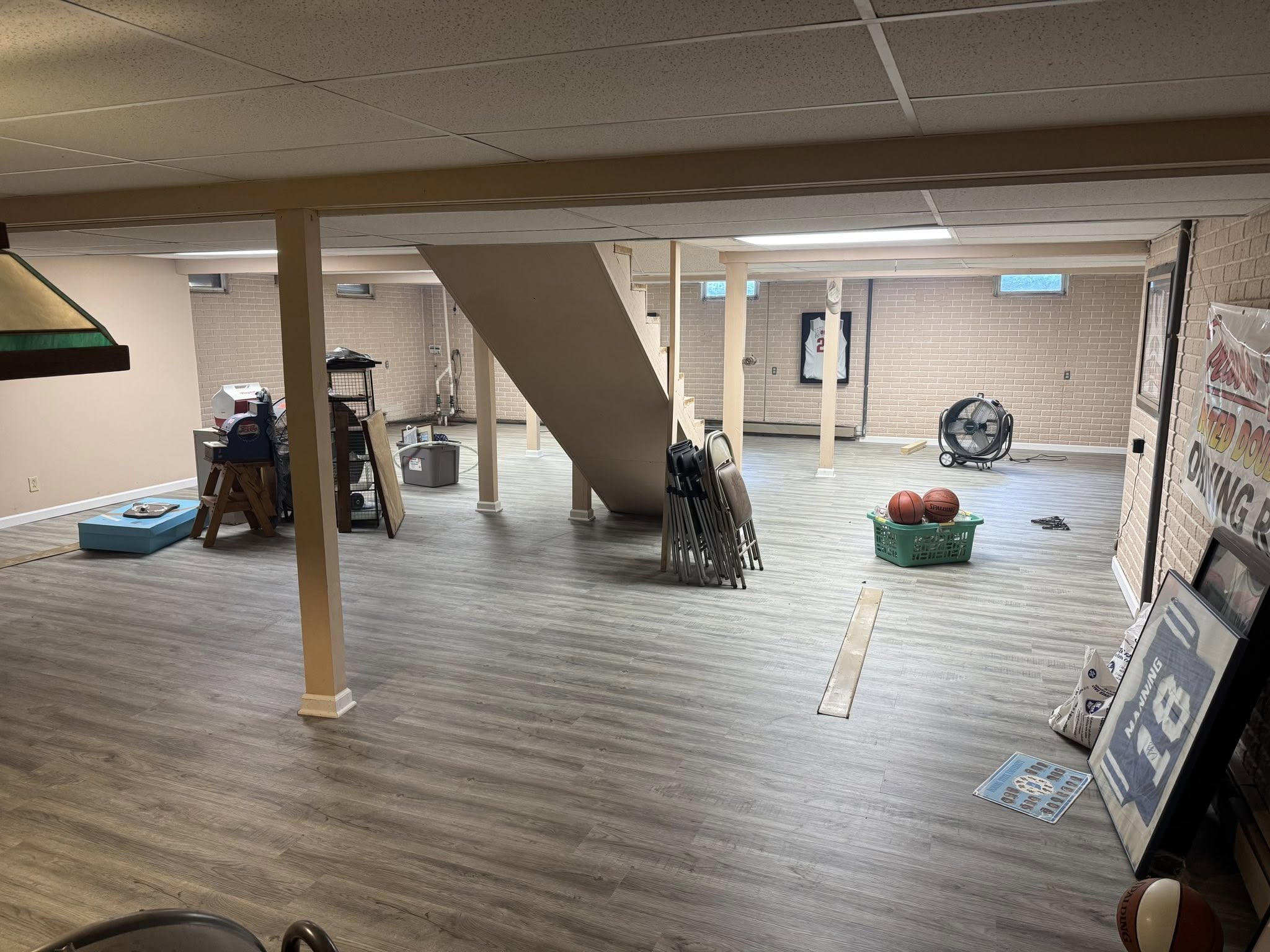 A spacious basement with laminate flooring, scattered sports equipment, folded chairs, gym equipment, and a staircase in the center.