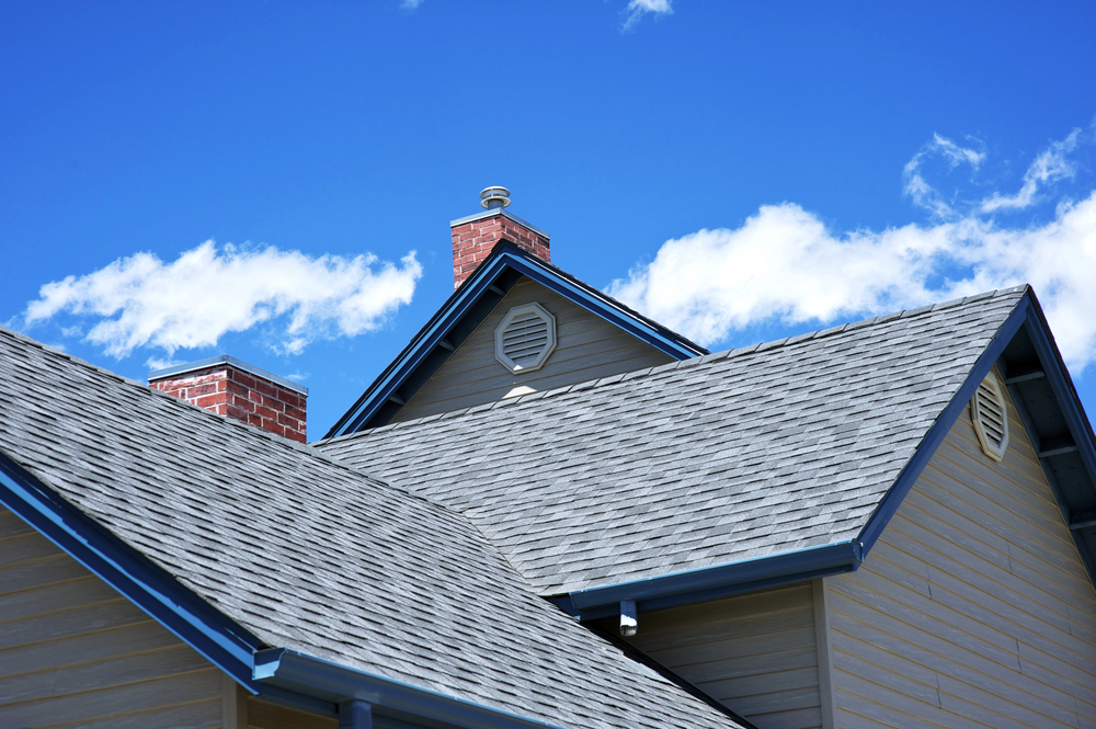 Close-up view of a house roof with gray shingles, brick chimneys, beige siding, and vents, under a blue sky with scattered white clouds.