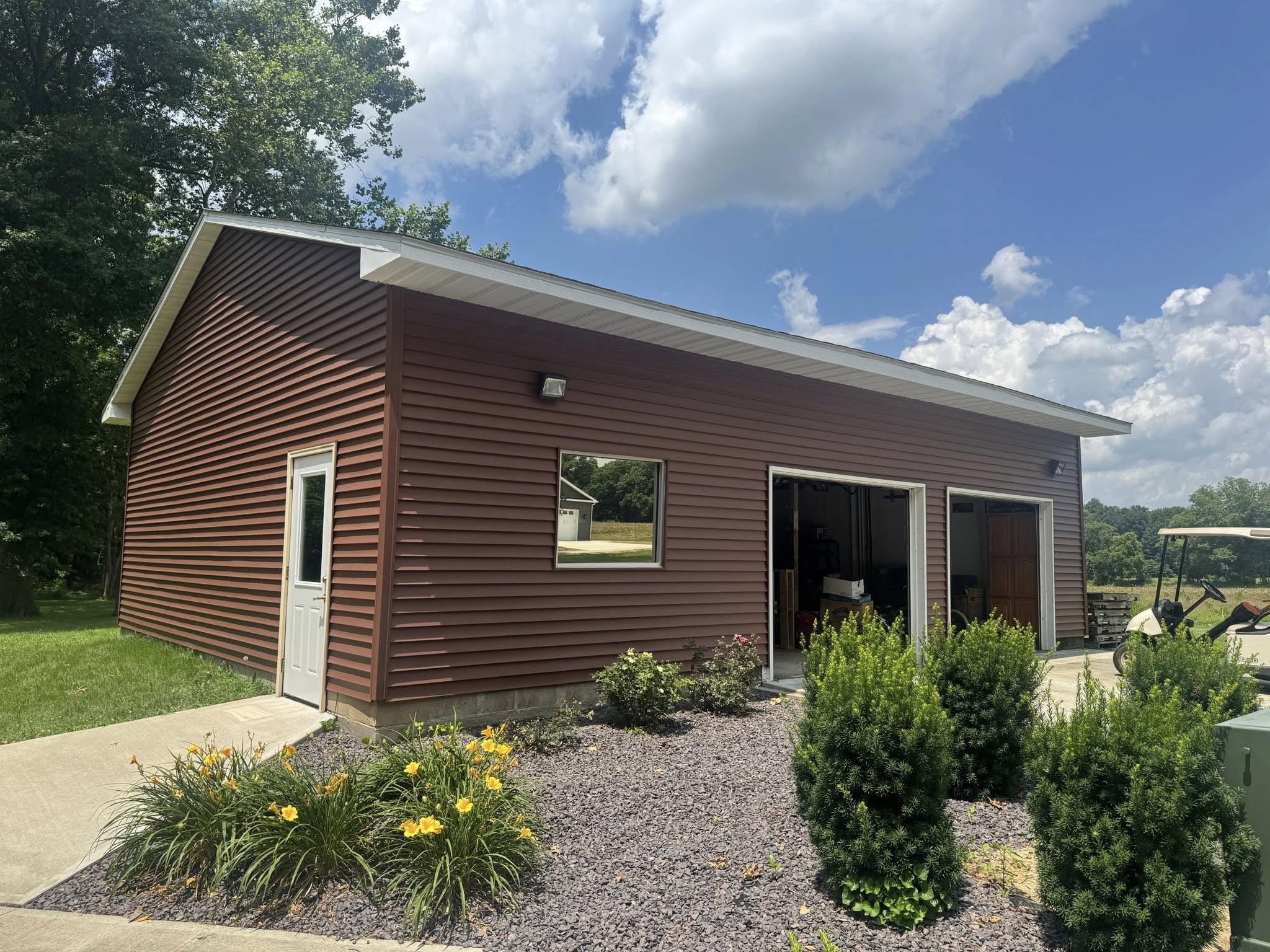 A brown metal garage with two open bays, a side door, and a window, surrounded by landscaping and shrubs under a partly cloudy sky.