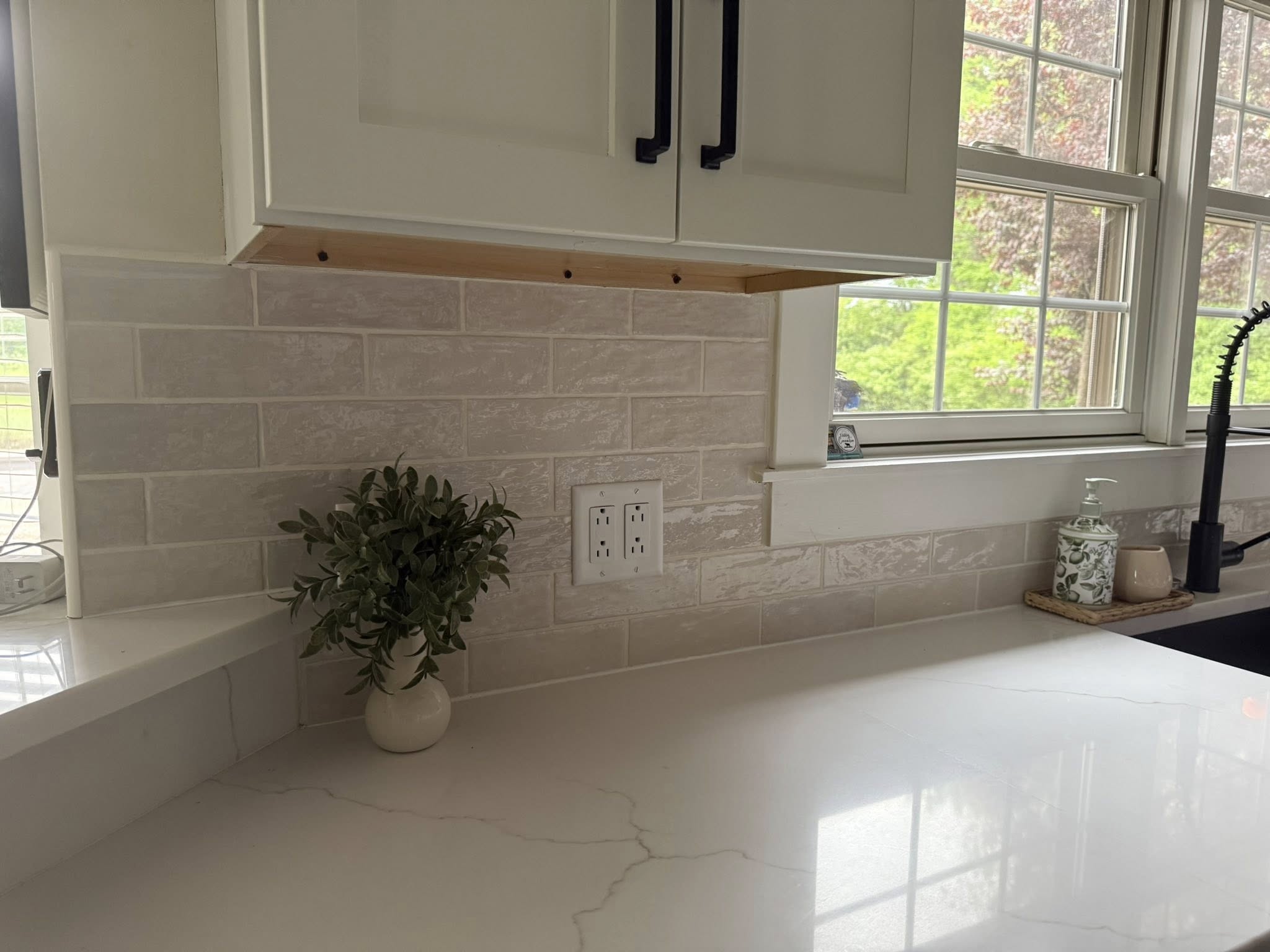 A clean kitchen counter with a small potted plant, white brick-style backsplash, electrical outlet, and a window above a black sink.