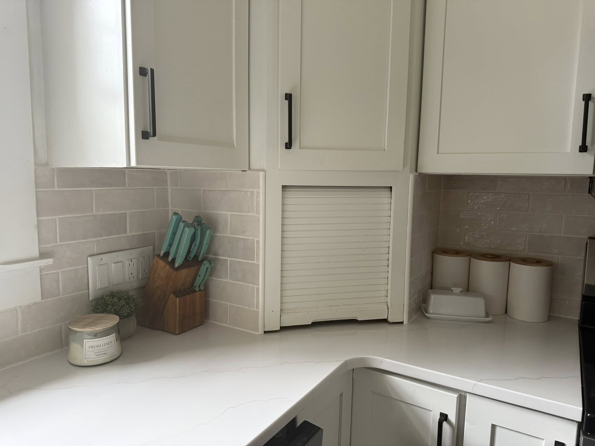A kitchen countertop with a knife block, three rolls of paper towels, a white container, a small plant, and a scented candle next to white cabinets and subway tile backsplash.